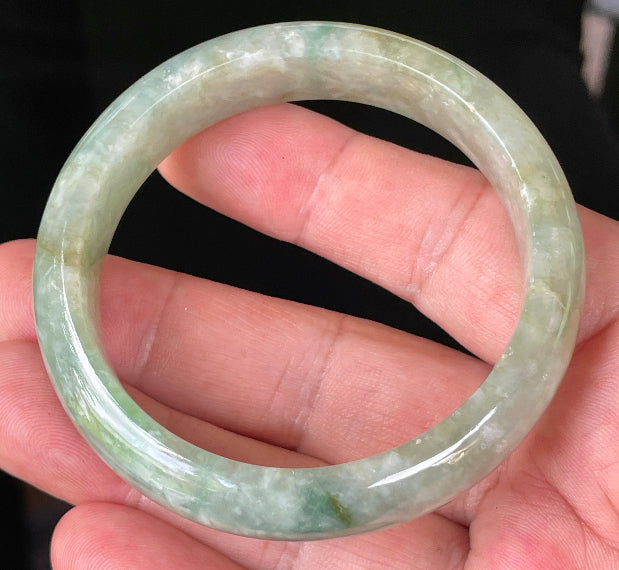 Hand holding a 55.4mm natural Grade A Burmese jadeite bangle in icy light apple and imperial green with milky white hues, item JB230