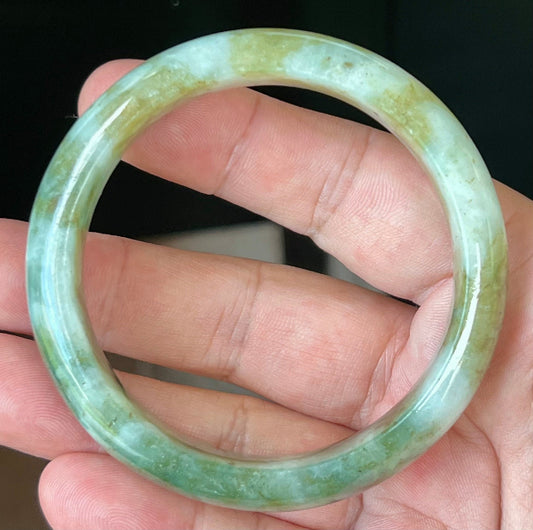 Hand holding a 58.8mm natural Grade A Burmese jadeite bangle in icy apple and moss green with milky white hues, item JB544