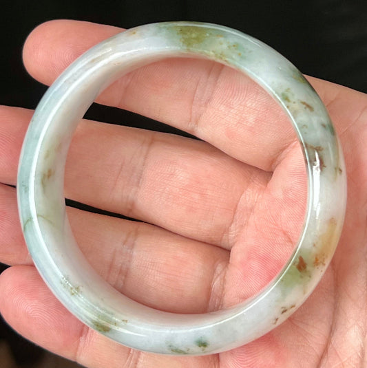 Hand holding a 57mm natural Grade A Burmese jadeite bangle in icy apple and moss green with milky white and red hues, item JB556
