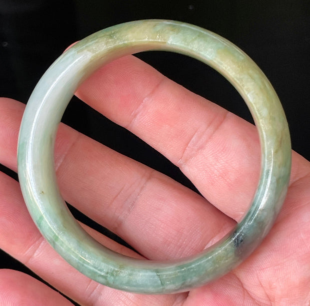 Hand holding a 55mm natural Grade A Burmese jadeite bangle in icy bluish apple green with honey and milky white hues, item JB233