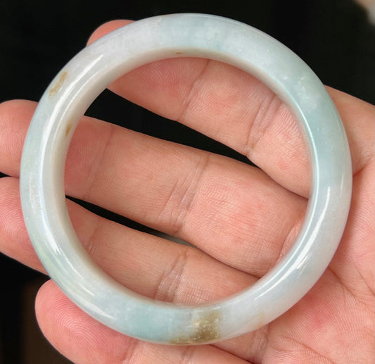 Hand holding a 53mm natural Grade A Burmese jadeite bangle in icy bluish milky white with honey brown hues, item JB519
