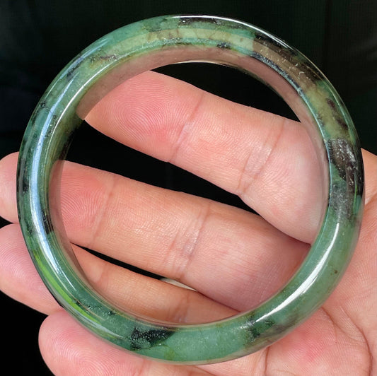 Hand holding a 58mm natural Grade A Burmese jadeite bangle in icy forest and apple green with black tones, item JB440