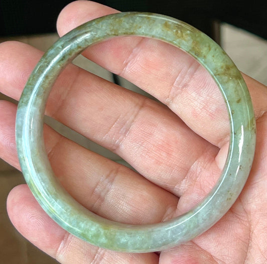 Hand holding a 58.8mm natural Grade A Burmese jadeite bangle in icy grayish milky white with forest green and honey hues, item JB512