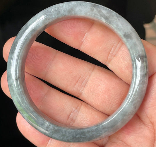 Hand holding a 57mm natural Grade A Burmese jadeite bangle in icy grey with misty milky white and honey hues, item JB364