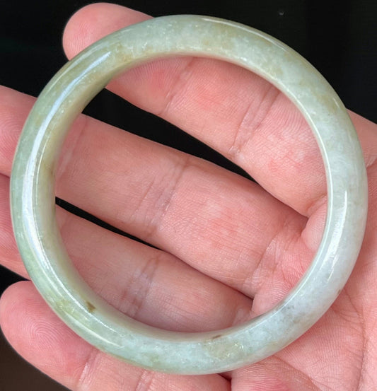 Hand holding a 56.7mm natural Grade A Burmese jadeite bangle in icy greyish milky white and apple green with honey hues, item JB538