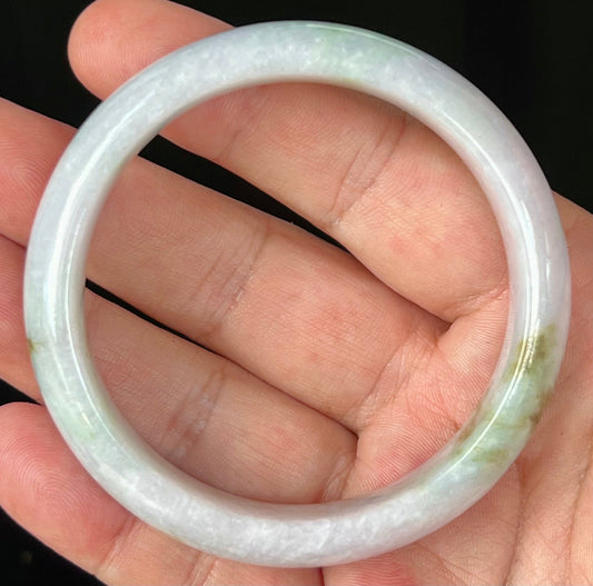 Hand holding a 58.2mm natural Grade A Burmese jadeite bangle in icy greyish milky white with apple green and honey hues, item JB534