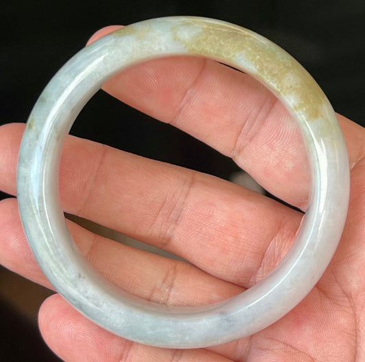 Hand holding a 59mm natural Grade A Burmese jadeite bangle in icy greyish milky white with moss green and honey hues, item JB546