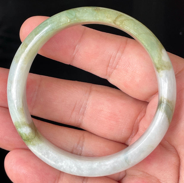 Hand holding a 57mm natural Grade A Burmese jadeite bangle in icy greyish milky white with apple green hues, item JB360