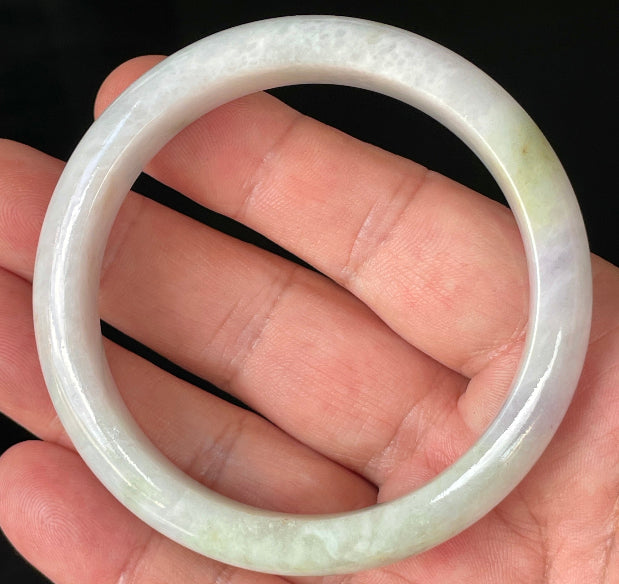 Hand holding a 58.9mm natural Grade A Burmese jadeite bangle in icy milky white with apple green hues, item JB235