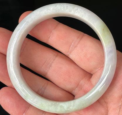 Hand holding a 58.9mm natural Grade A Burmese jadeite bangle in icy milky white with apple green hues, item JB235