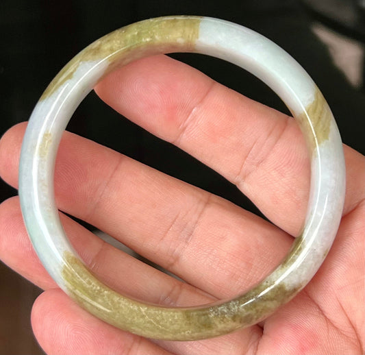 Hand holding a 58.5mm natural Grade A Burmese jadeite bangle in icy milky white with golden brown and moss green, item JB555